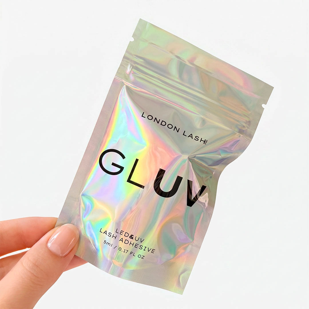 A hand holding a holographic silver resealable pouch of London Lash Gluv LED&UV Lash Adhesive against a clean white background.