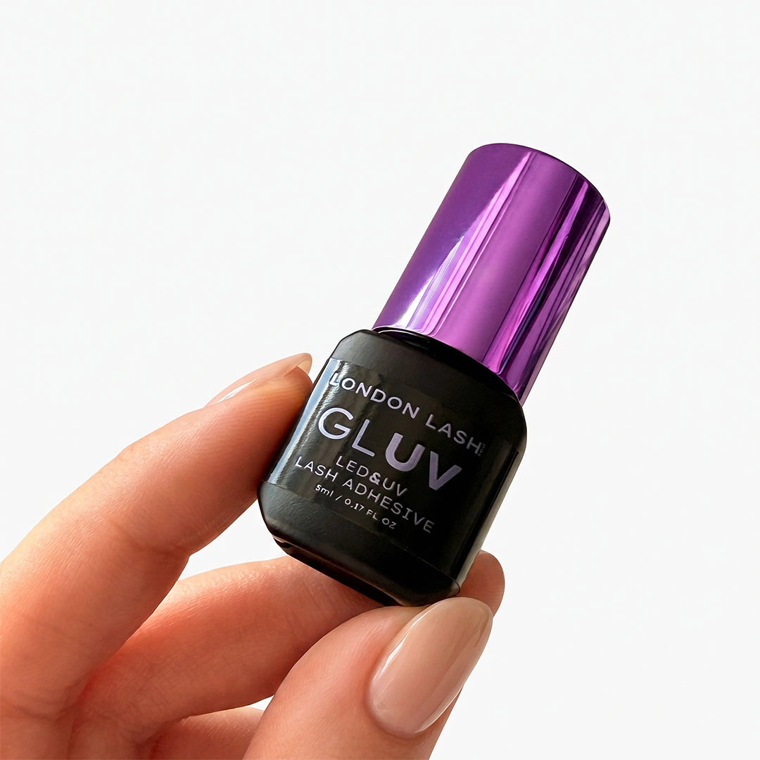 A hand holding a bottle of London Lash Gluv LED & UV Lash Adhesive. The bottle is black with a metallic purple cap, set against a clean white background.