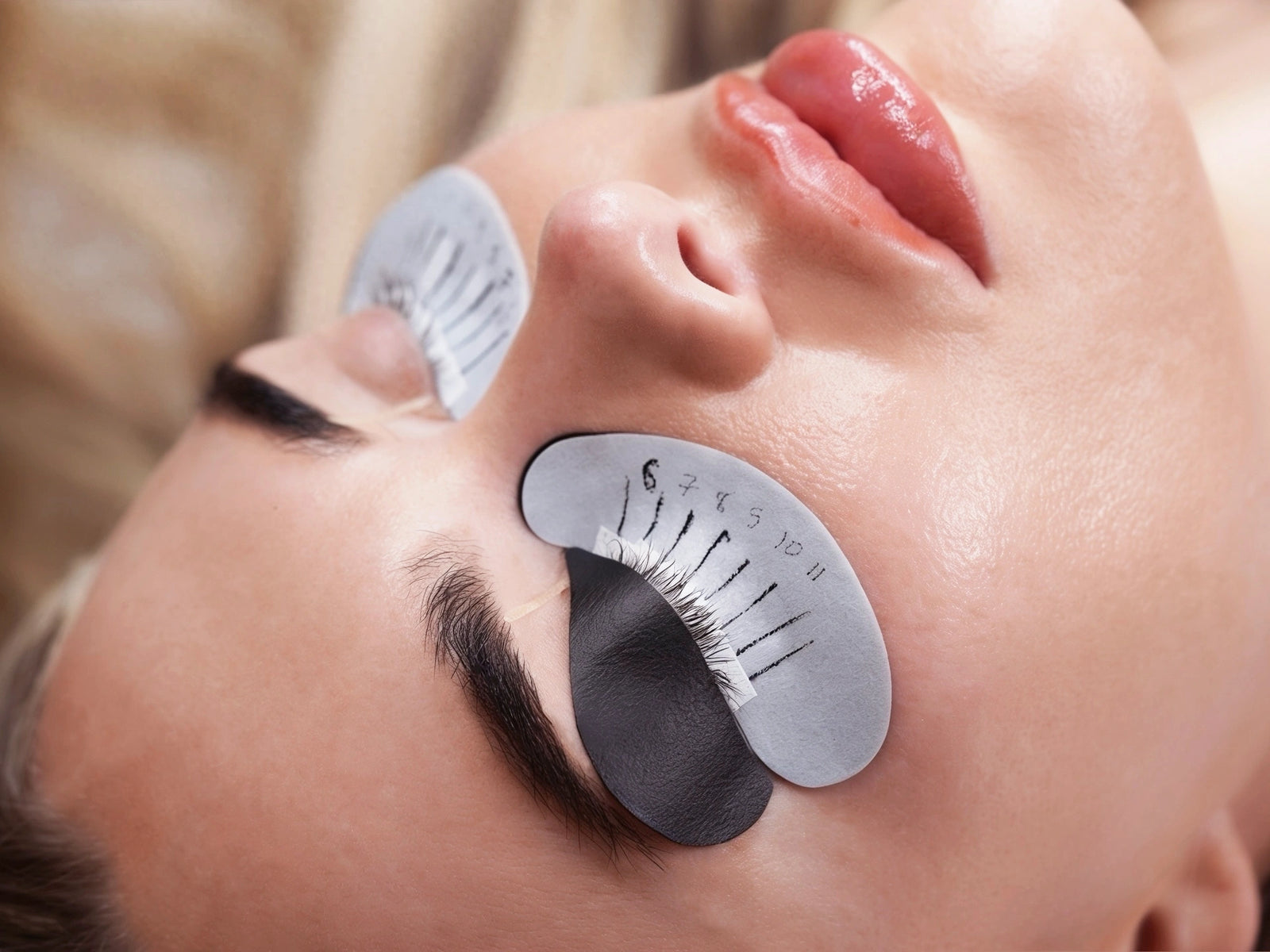 Close-up of a person lying down with false eyelashes applied under closed eyes using a measurement guide, with additional lash pads visible.